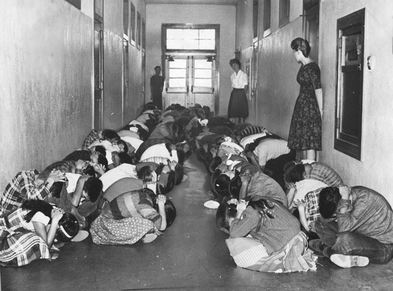 Take Cover drill, Elysian Heights School, 1982.  Courtesy Los Angeles Public Library.  Shows children sitting in a school hallway, with heads ducked, and covering heads with hands.